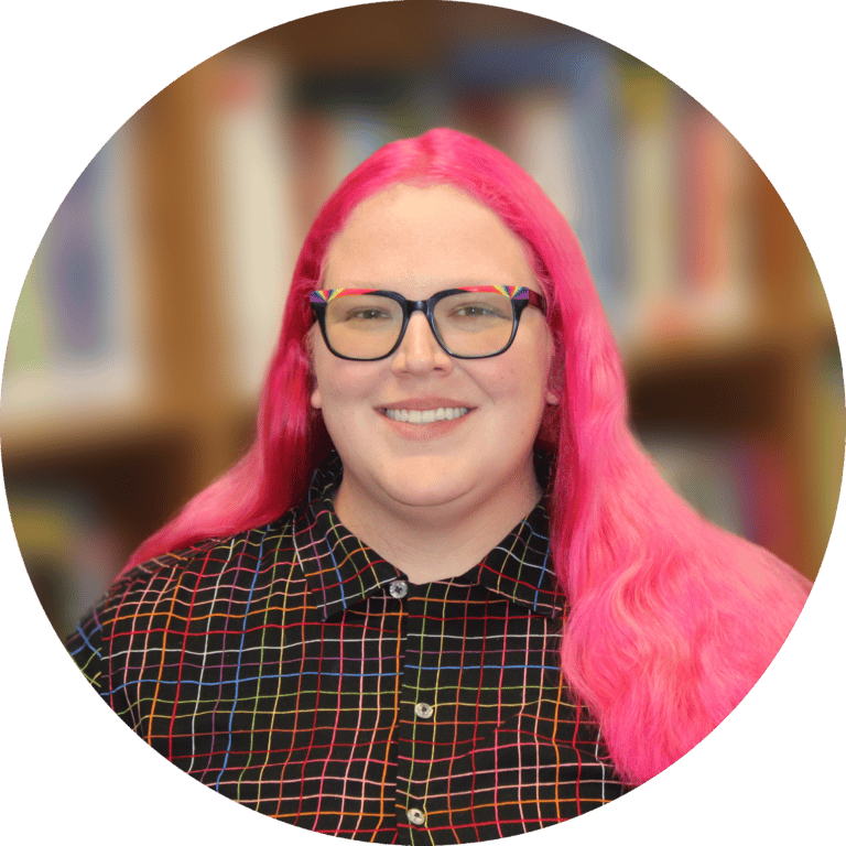 a white non-binary person with shoulder length vivid pink hair smiles at the camera. They are wearing black rimmed glasses with rainbow accents and a black and rainbow plaid short sleeve button down shirt.